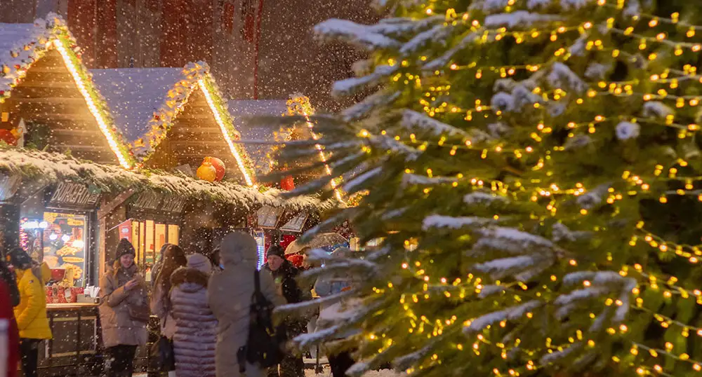 People peruse booths at a Christmas market with a big, lit-up evergreen tree in the foreground