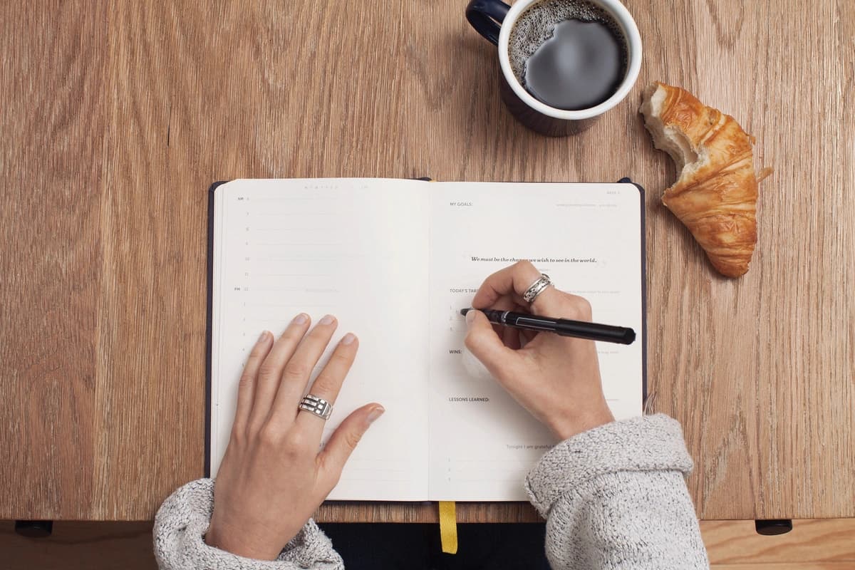 Person writes in a notebook at a table, with croissant and coffee